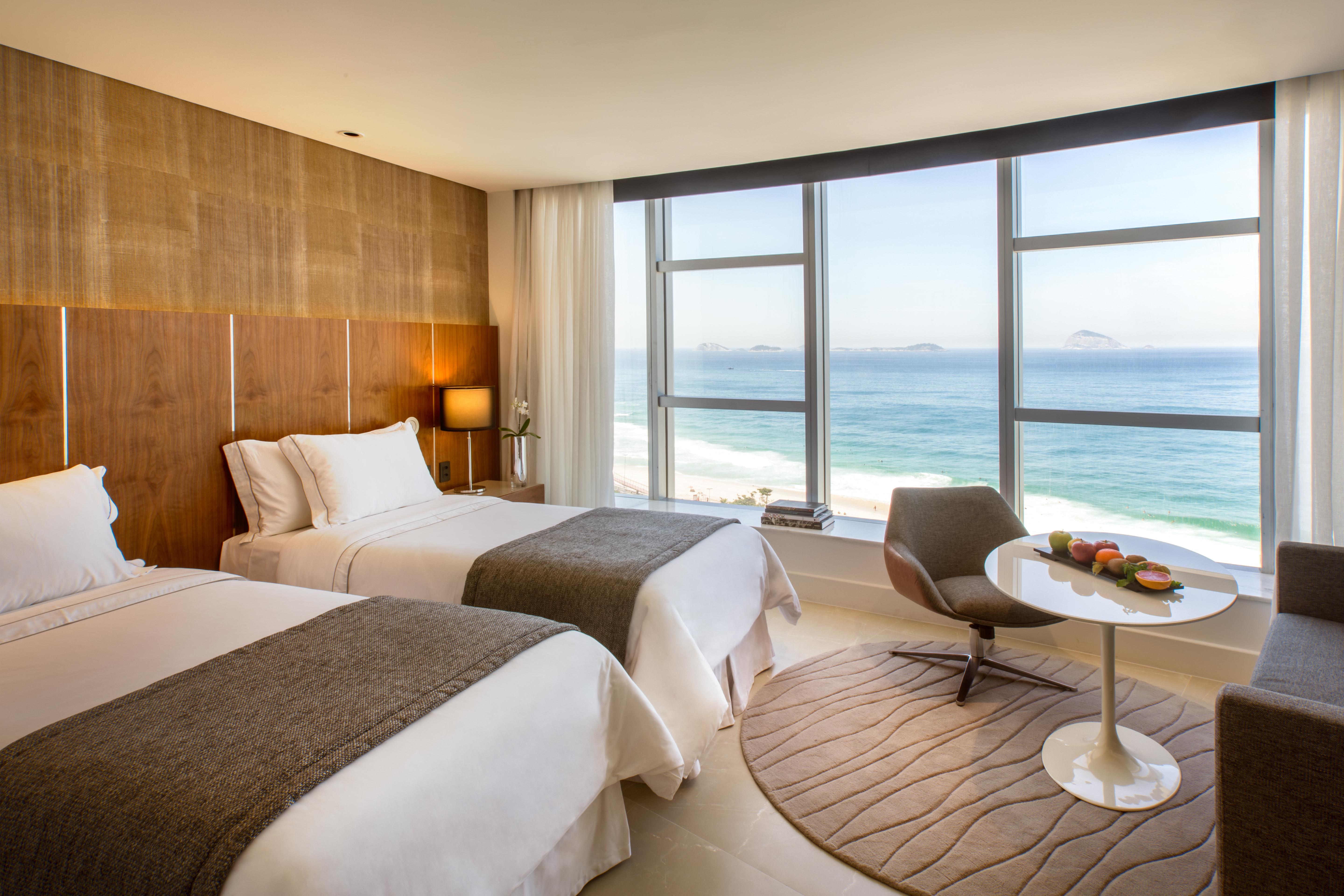 Hotel Nacional Rio De Janeiro - Oficial 외부 사진 The photo shows a modern hotel room with two beds adorned in white linens and gray throws. Large windows allow natural light to flood the space, offering a stunning view of the ocean. In the corner, there's a comfortable chair and a small round table